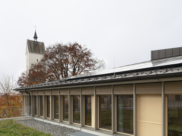 Modernes Flachdachgebäude mit Naturumgebung Modernes Flachdachgebäude mit großer Fensterfront und Holzfassade, umgeben von Grünfläche; im Hintergrund ein Kirchturm und herbstliche Bäume.