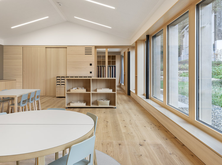 Moderner Gemeinschaftsraum mit Fensterfront Heller Gemeinschaftsraum im Kindergarten St. Vincenz mit runden Tischen, Holzfußboden und großer Fensterfront mit Blick ins Grüne.