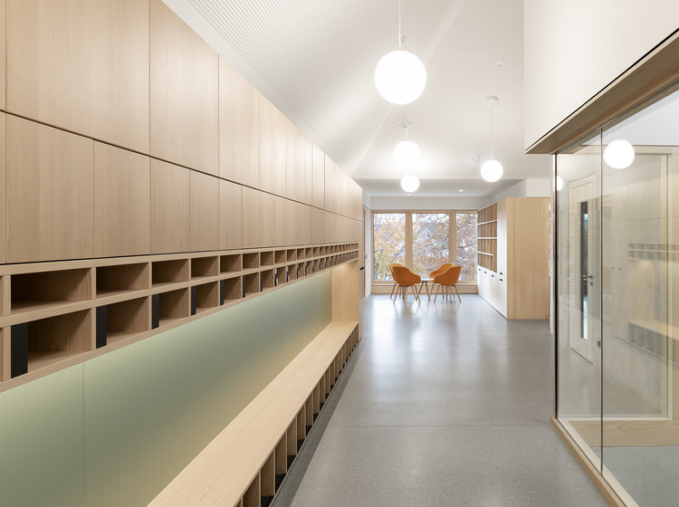 Moderner Flurbereich mit Stauraum und Sitzgelegenheit Heller Flur mit Holzregalen und Sitzbank, modernen Glastrennwänden und runden Deckenleuchten; im Hintergrund zwei orangefarbene Stühle.