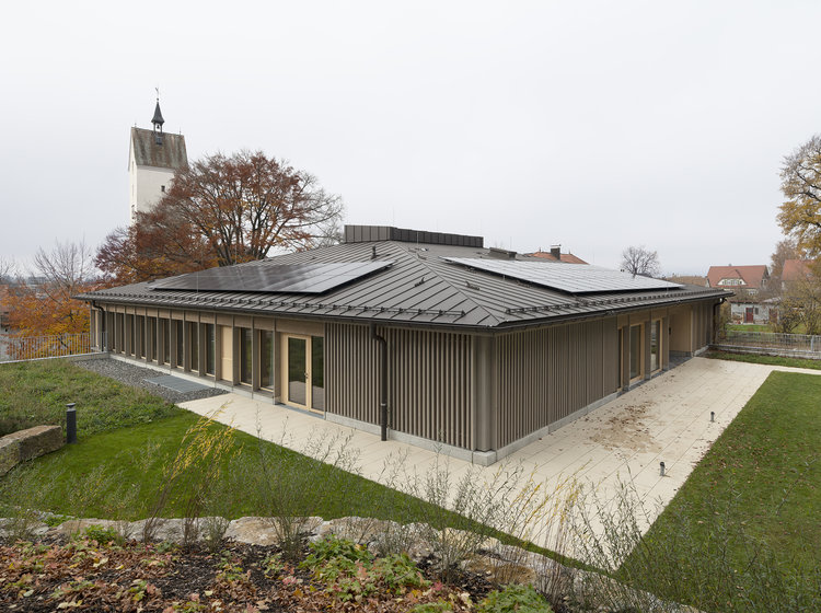 Nachhaltiger Neubau mit Solarpanels und Holzfassade. Energieeffizienter Neubau mit dunkler Holzfassade, Flachdach und Solarpanels; umgeben von Rasenflächen, im Hintergrund ein Kirchturm und Bäume.